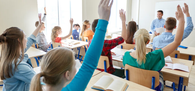 Ungdomar i ett klassrum som håller upp en arm. Läraren står framför klassen.