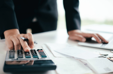 Person som står vid ett skrivbord. Har papper på ena sidan och har handen på en miniräknare på andra sidan.