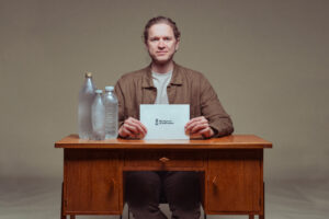 Man som sitter vid ett skrivbord, han håller ett papper med texten vatten. På skrivbordet står vattenflaskor.