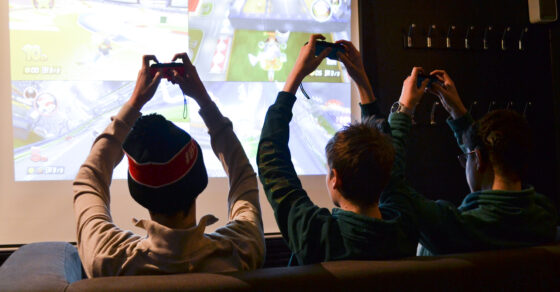 Tre ungdomar som sitter framför en skärm. Håller upp sina händer hållandes spelkonsoller.