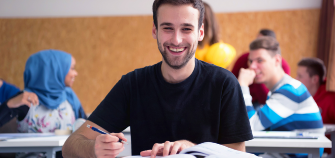 En kille sitter i ett klassrum och studerar. Bakom honom sitter klasskamreaterna och diskuterar.