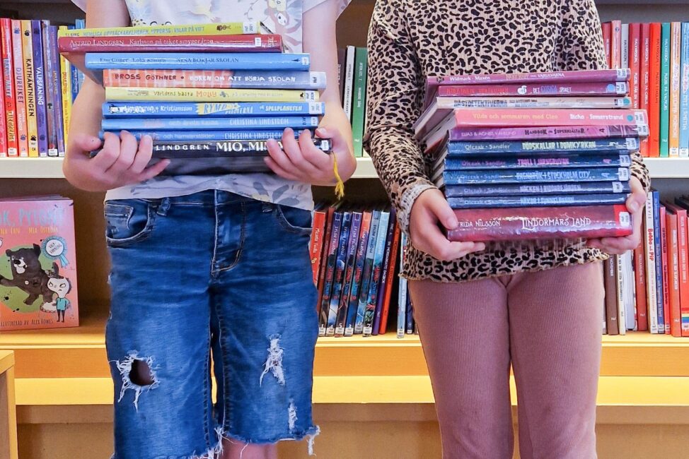 Två barn som står framför en bokhylla med böcker. I famnen håller de en varsin hög med böcker.